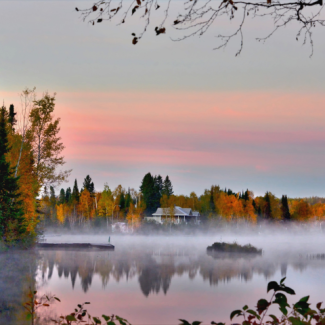 Fog hovering over a lake with a backdrop of autumn trees, houses, and the sunrise.