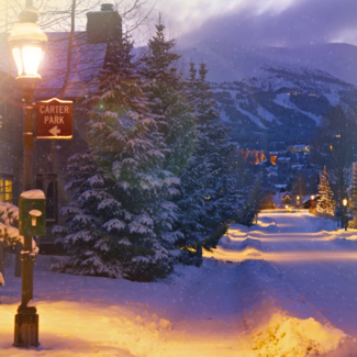 A snowy city with mountains and Christmas lights in the background and a light pole in the forefront with a sign reading “Carter Park”.